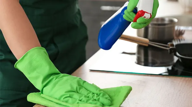 Person wearing green gloves sanitizing a kitchen counter with a spray bottle and cloth