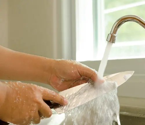 Hands rinsing a chef's knife under warm running water in a kitchen sink