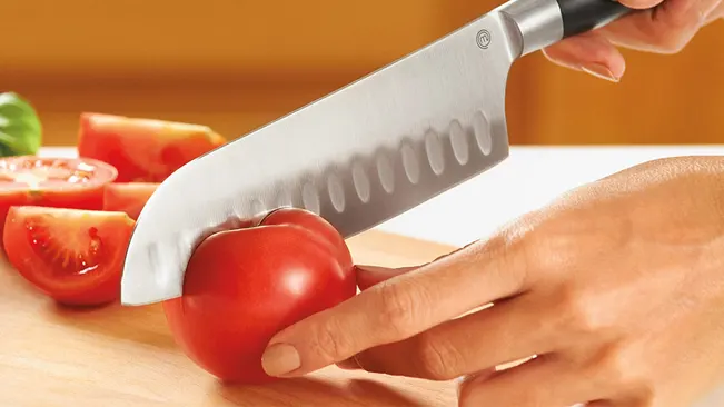 Close-up of hands skillfully slicing a ripe tomato with a Santoku knife on a wooden cutting board