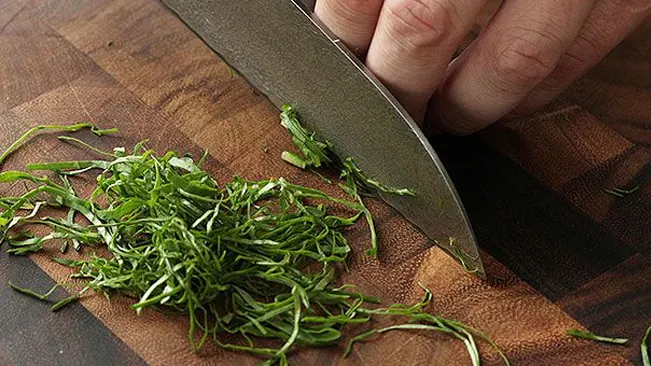 Hand using a Santoku knife to chop green herbs on a wooden cutting board