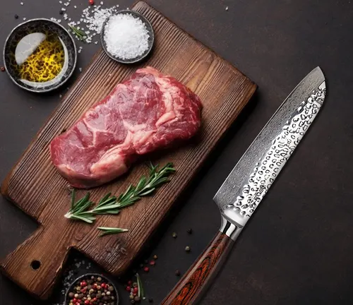 Santoku knife next to a raw steak, herbs, and seasonings on a wooden cutting board