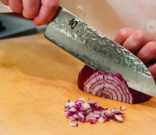 Hand holding a Santoku knife dicing a red onion on a wooden cutting board