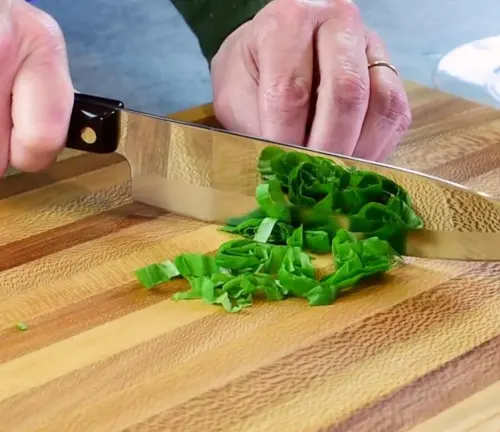 Person using a Santoku knife to chiffonade green leafy vegetables on a wooden cutting board