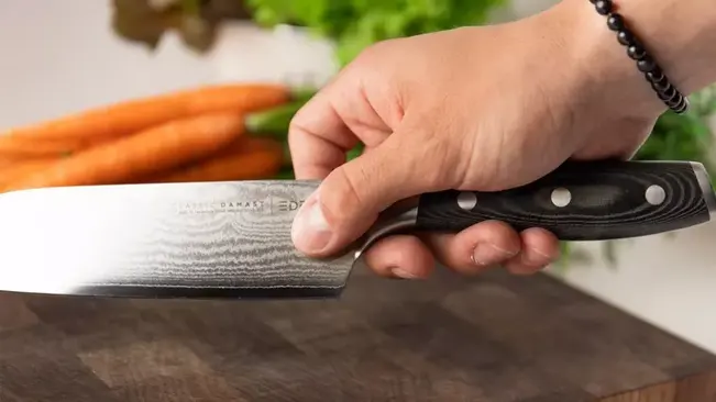 Close-up of a person’s hand demonstrating the proper grip on a Santoku knife, with carrots and parsley in the background